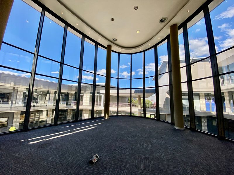 Spacious, modern office area featuring expansive floor-to-ceiling windows, high ceilings with recessed lighting, and neutral carpet tiles, offering abundant natural light.