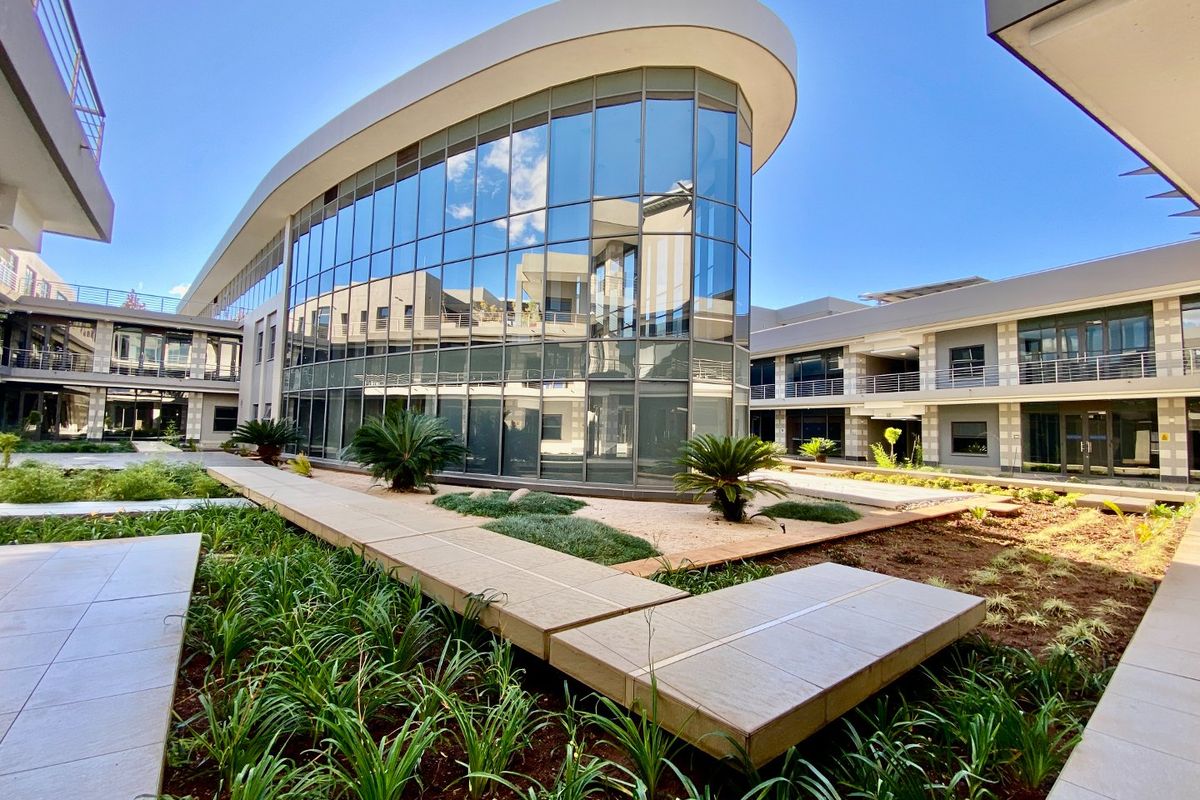 Modern commercial office building featuring a curved glass facade, elevated concrete walkways, and well-maintained landscaped garden areas.