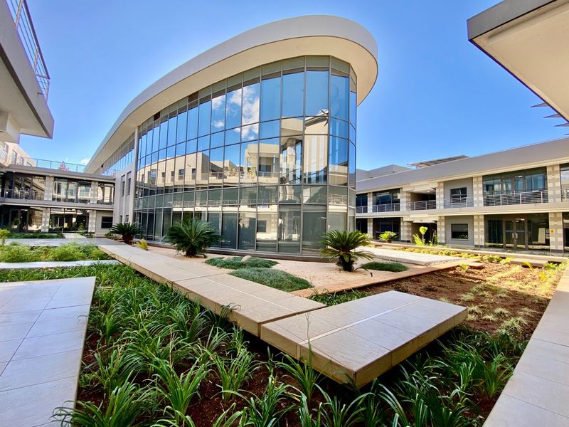 Modern commercial office building featuring a curved glass facade, elevated concrete walkways, and well-maintained landscaped garden areas.