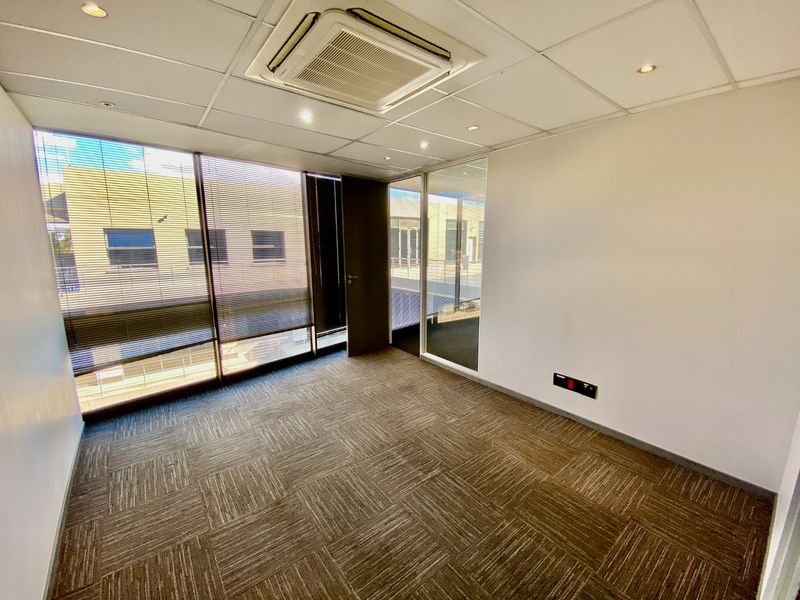 A bright, vacant commercial office space featuring carpet tiles, a suspended ceiling with integrated air conditioning, and large windows with venetian blinds.