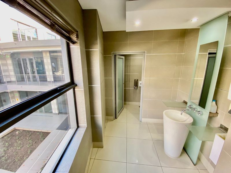 A modern commercial bathroom featuring a sleek pedestal basin, tiled finishes, and large windows overlooking the building courtyard.