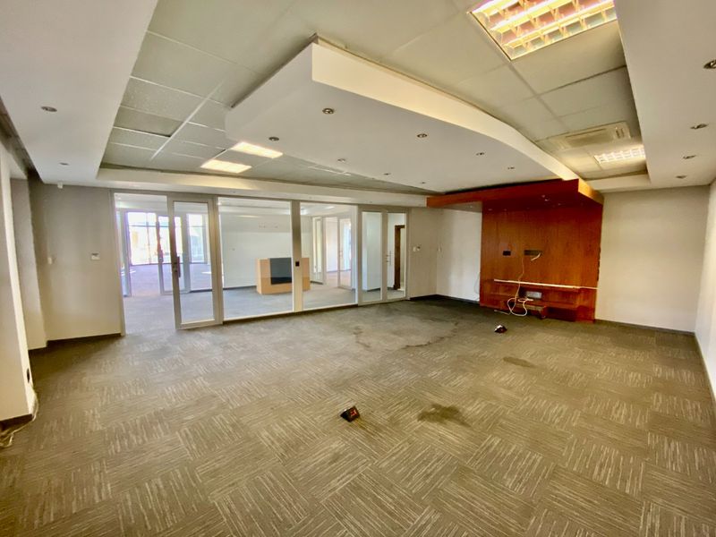 An empty commercial office space featuring glass partitions, a suspended ceiling with recessed lighting, and a built-in wooden media console.