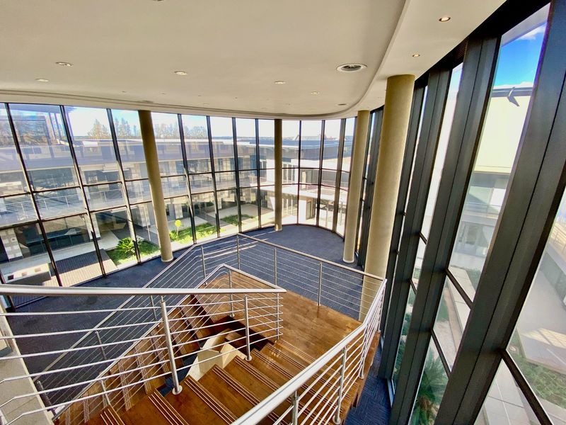 Spacious commercial atrium featuring a wooden staircase with stainless steel railings, surrounded by extensive floor-to-ceiling glass windows overlooking the courtyard.