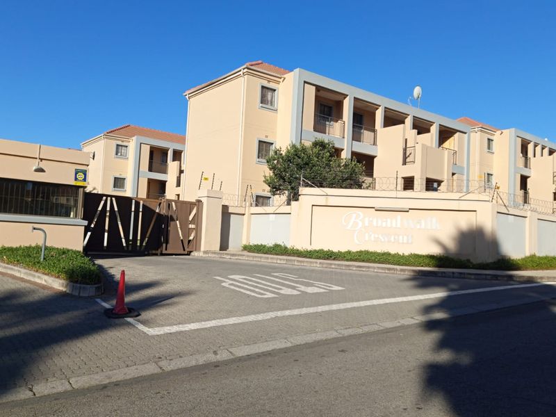 Front entrance view of Broadwalk Crescent apartment complex, featuring secure gated access, perimeter wall with electric fencing, and multi-story residential buildings.