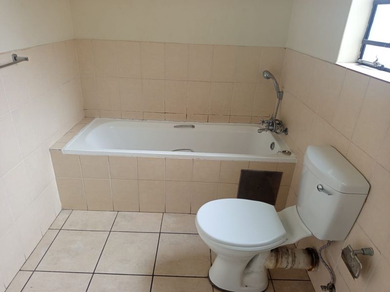 A dated bathroom featuring a built-in bathtub with shower mixer and a standard toilet, finished with neutral wall and floor tiles.