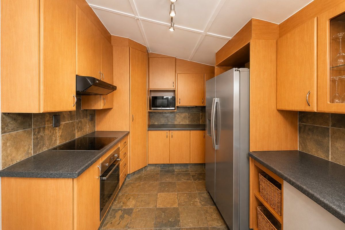 A functional kitchen featuring wood-tone cabinetry, dark laminate countertops, slate-style floor tiles, and integrated appliances including an oven, cooktop, and microwave.