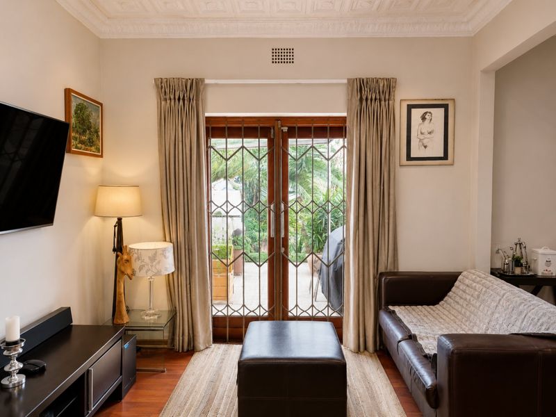 A cozy living room featuring timber flooring, ornate ceiling detailing, and double glass doors with security gates leading to an outdoor patio area.