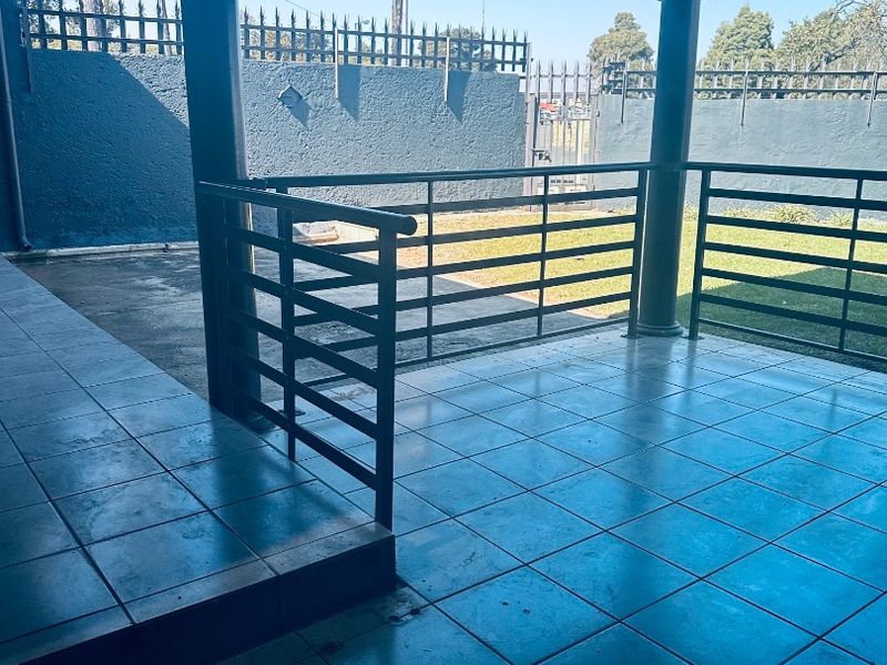 A covered outdoor patio area featuring tiled flooring, metal railings, and support columns, overlooking a private garden space with perimeter walling.