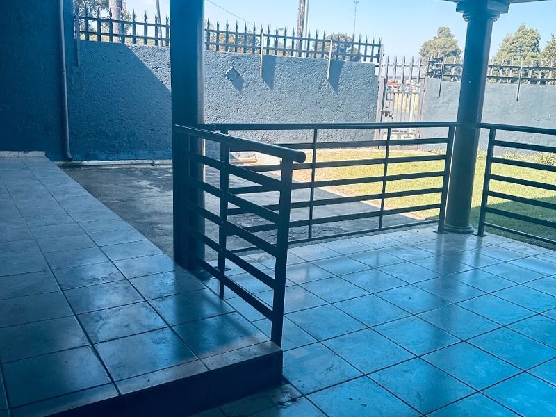 A covered outdoor patio area featuring tiled flooring, metal railings, and support columns, overlooking a fenced garden space.