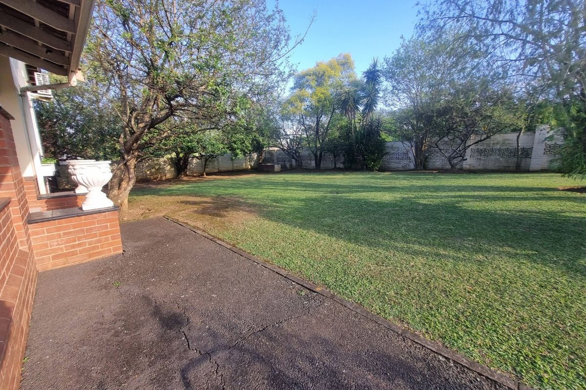 A spacious rear garden featuring a large lawn area, mature trees, and a paved patio space enclosed by a brick boundary wall.