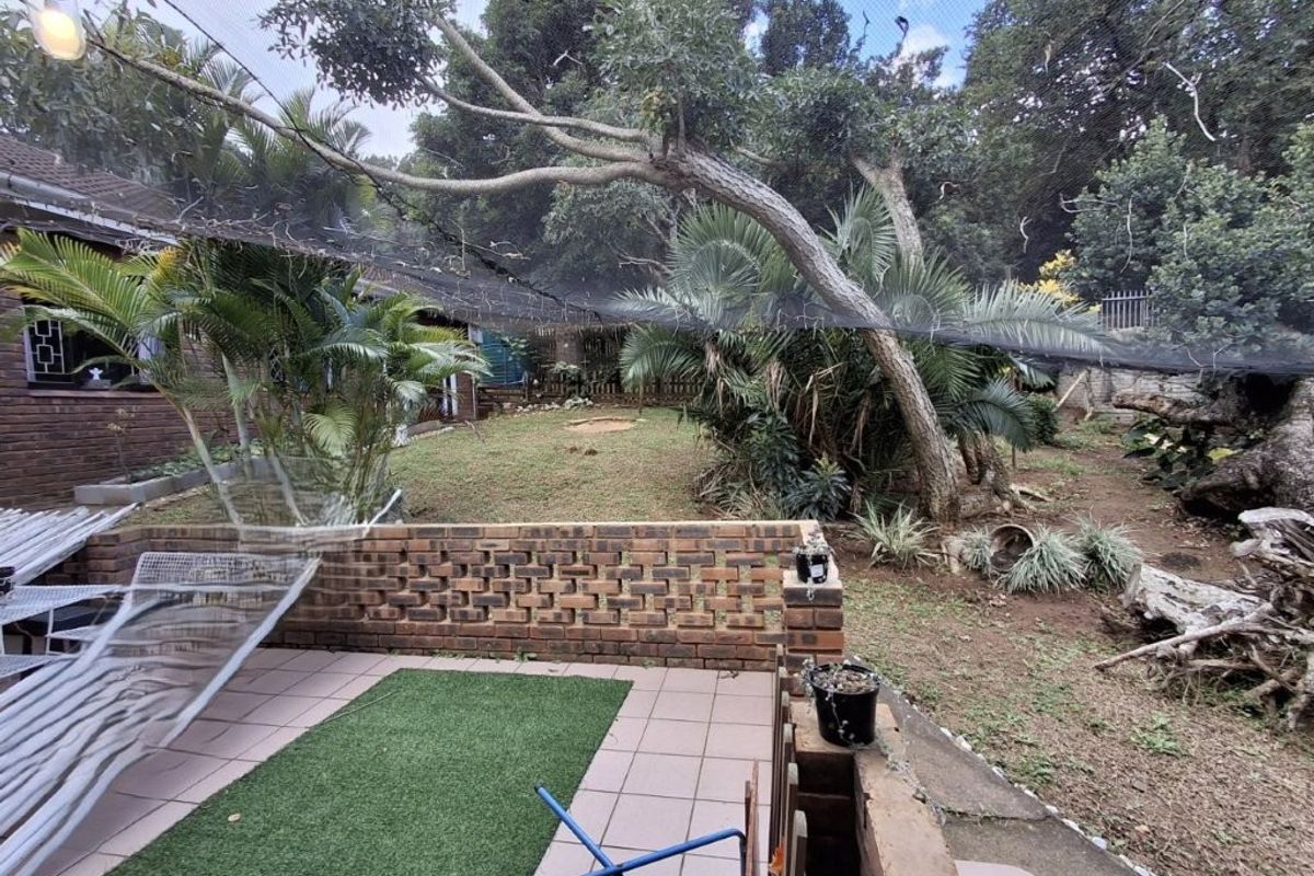 A rear garden area featuring a tiled patio, a low brick retaining wall, and established trees covered by overhead netting.