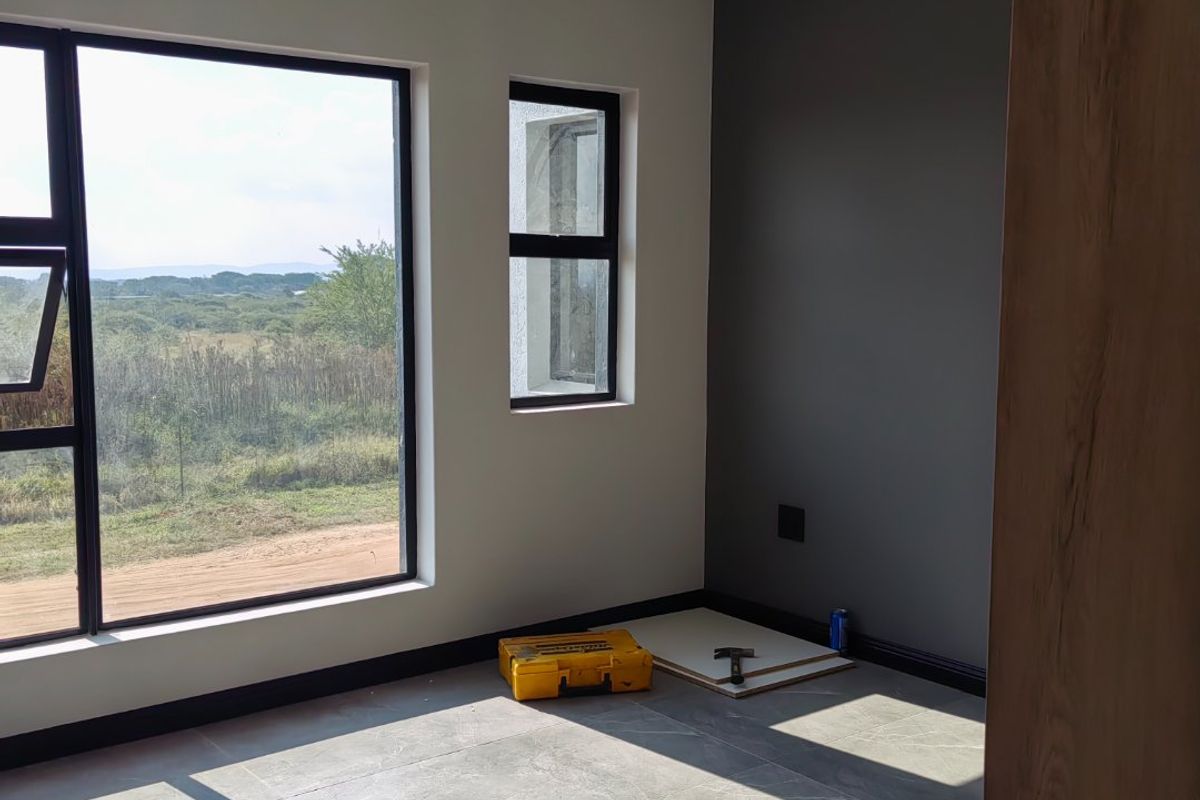 A bright, modern bedroom featuring large windows with natural views, neutral tiled flooring, and a dark grey accent wall.
