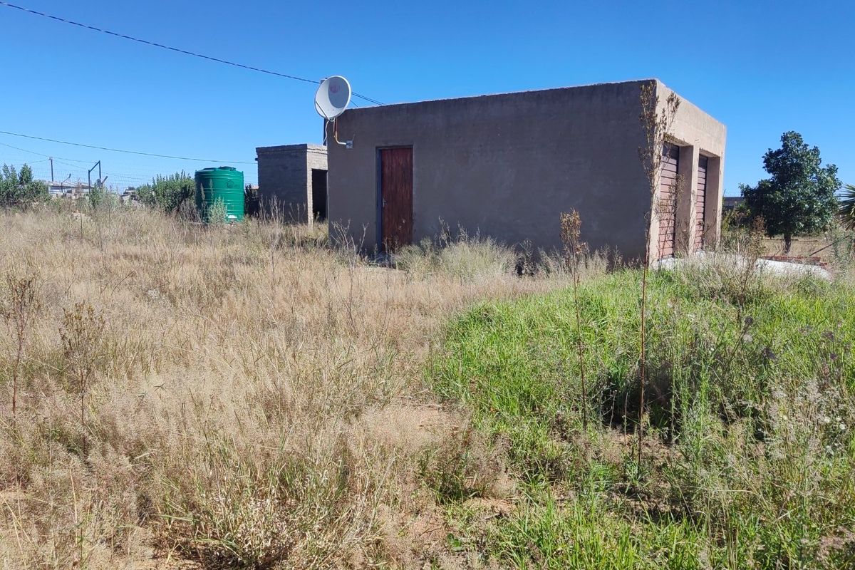 Exterior view of a detached garage structure with an adjacent water storage tank, set on a large plot with overgrown vegetation.