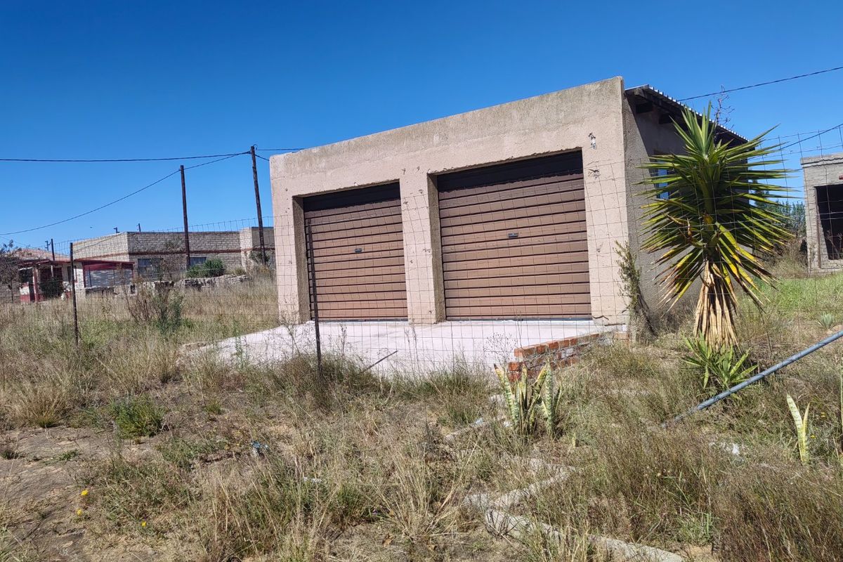A detached double garage structure with roller doors, situated on a large, overgrown plot of land.