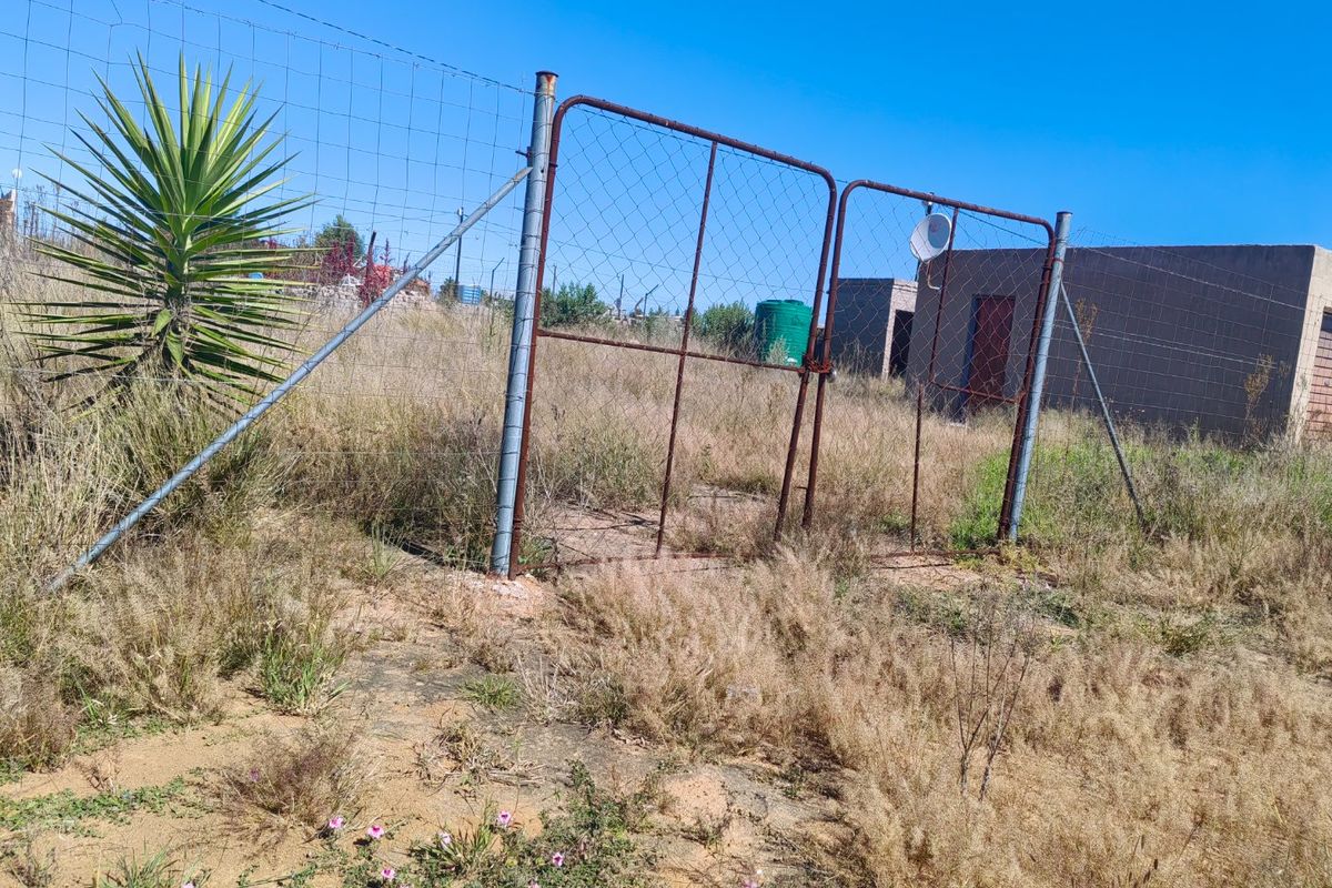 An outdoor area featuring a wire mesh fence, a metal gate, and a water tank, set within an overgrown, unmaintained plot of land.