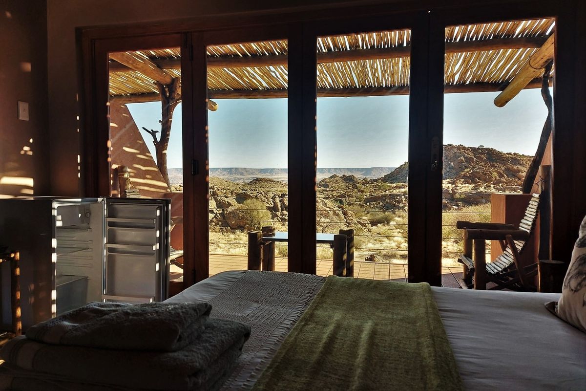 A bedroom featuring large timber-framed glass doors that open onto a private deck with a thatched roof and expansive desert landscape views.