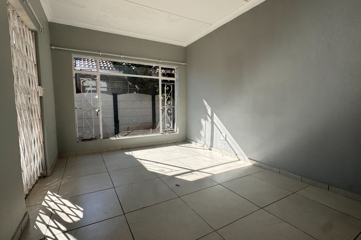 A bright, empty living room featuring neutral tiled flooring, large windows with security bars, and a secure entrance gate.