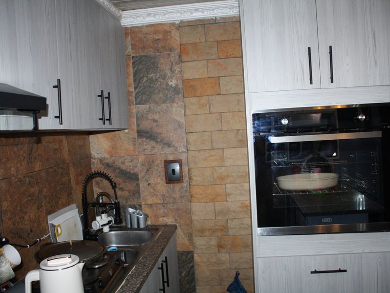 A compact kitchen area featuring light wood-grain cabinetry, a built-in oven, extractor fan, and stone-look wall tiling.