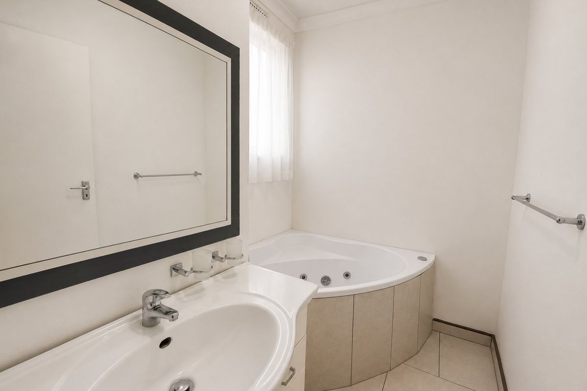 A clean and functional bathroom featuring a corner spa bath, vanity basin, and neutral tiled flooring, offering a bright and well-maintained space.