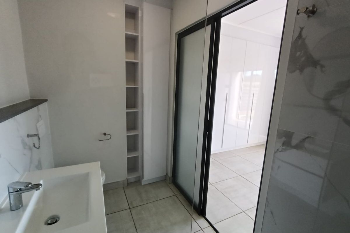 Modern bathroom featuring a sleek vanity, built-in shelving, and a glass-panel door leading to an adjacent bedroom with fitted wardrobes.