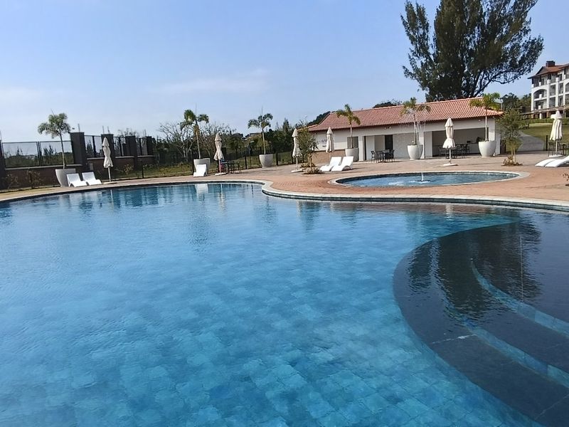 A well-maintained communal swimming pool area featuring paved surrounds, landscaped grounds, and a clubhouse building within a residential apartment complex.