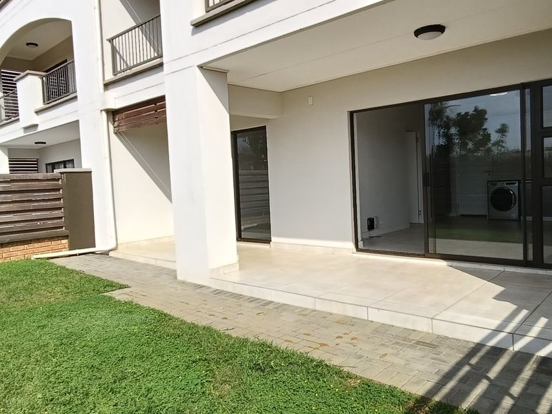 A ground floor apartment featuring a covered tiled patio, brick paved walkway, and a private garden area enclosed by timber fencing.