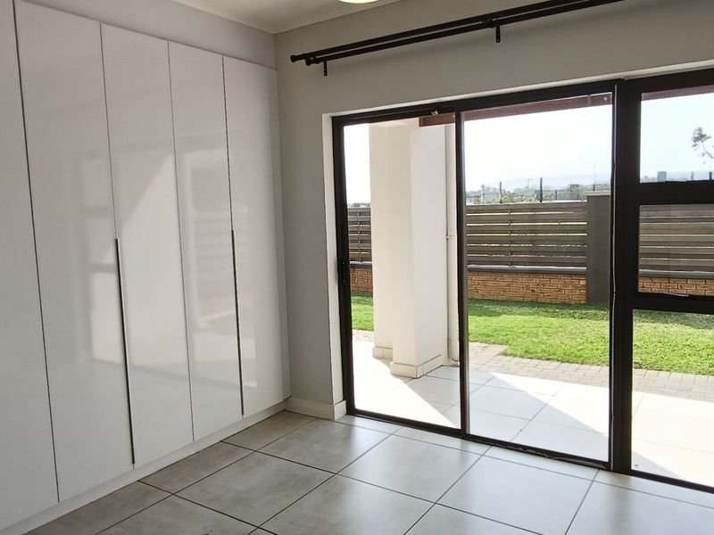 A bright, empty bedroom featuring modern white built-in cupboards, tiled flooring, and a sliding glass door providing direct access to the patio and garden.