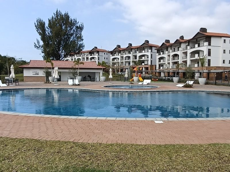 A well-maintained communal swimming pool area featuring a paved deck, surrounding landscaped grounds, and views of the apartment complex buildings.