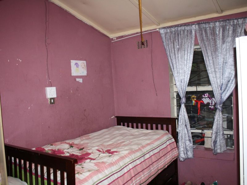A bedroom featuring purple walls, security bars on the window, and basic electrical fixtures. The room requires cosmetic updates and wall repairs.