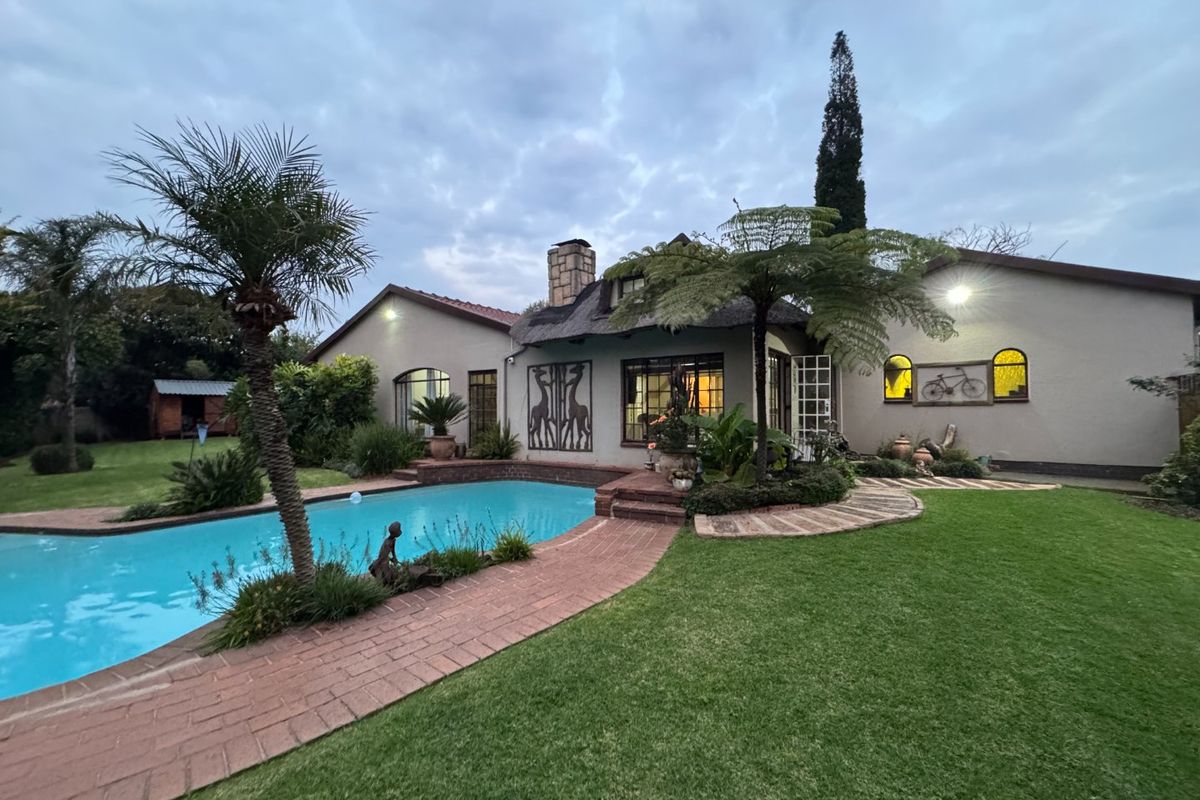 A well-maintained rear garden featuring a swimming pool, brick pathways, and a charming house with a thatched roof and stone chimney.