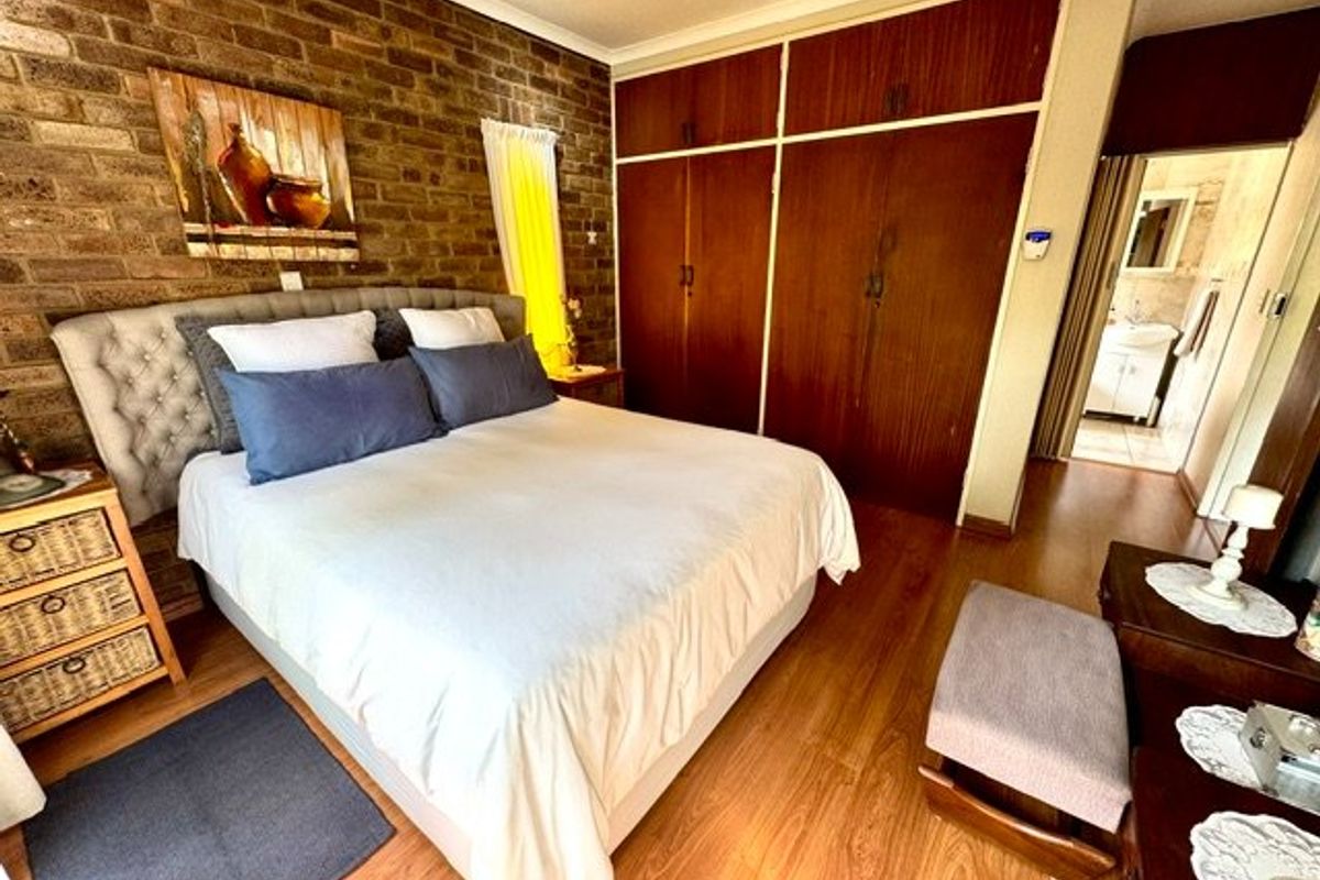 A bedroom featuring an exposed brick feature wall, laminate flooring, and built-in wooden cupboards, with an adjoining bathroom visible in the background.