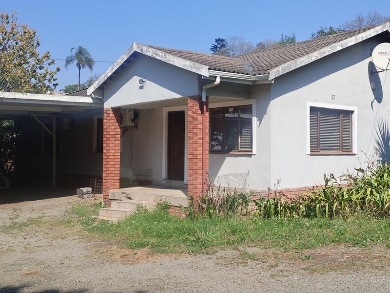 Front view of a single-story house featuring a carport, brick-accented entrance, and tiled roof, set on a gravel driveway with surrounding garden area.
