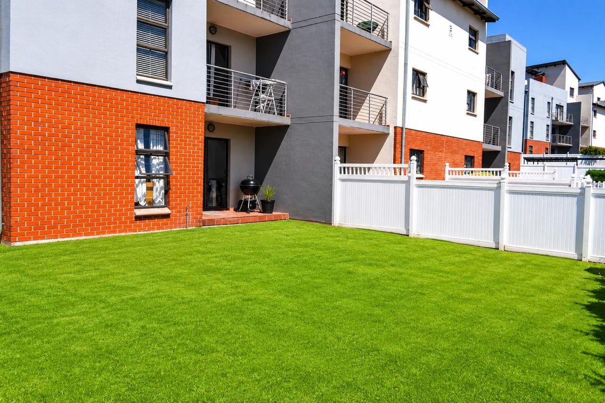 A well-maintained rear garden area featuring a manicured lawn, enclosed by white vinyl fencing, adjacent to a modern apartment building with brick and render facade.