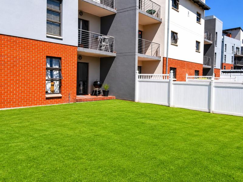 A well-maintained rear garden area featuring a manicured lawn, enclosed by white vinyl fencing, adjacent to a modern apartment building with brick and render facade.