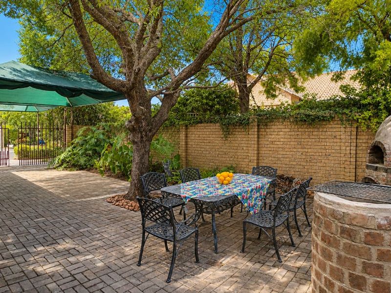 A paved outdoor patio area featuring a built-in pizza oven, brick braai, and established trees, enclosed by a brick boundary wall.