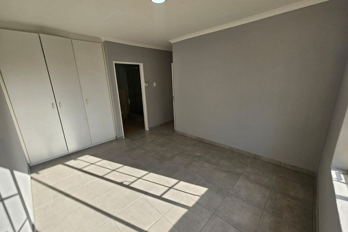 A bright, empty bedroom featuring neutral grey walls, tiled flooring, and built-in white cabinetry, with an adjoining doorway leading to an en-suite bathroom.