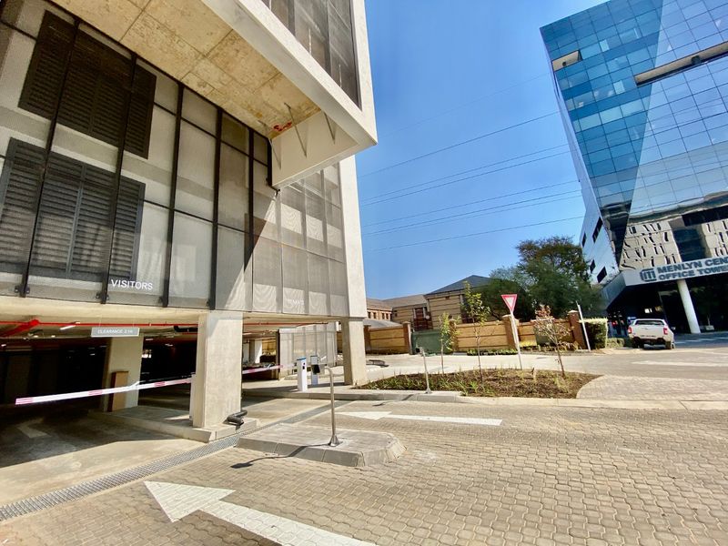 Exterior view of a commercial parking facility entrance featuring a paved driveway, boom gate access, and modern concrete and metal mesh architectural elements.