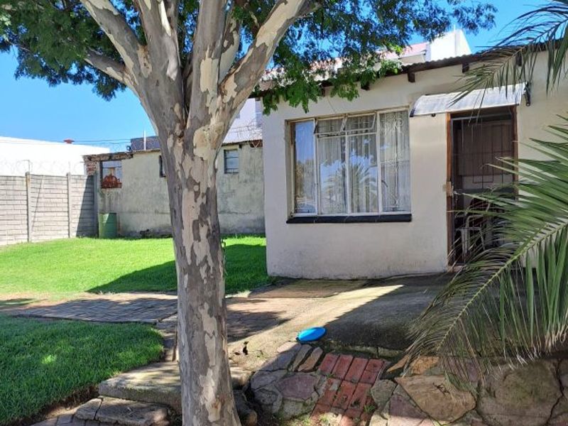 Rear exterior view of the property featuring a garden, mature tree, stone retaining wall, and paved patio area leading to the entrance.