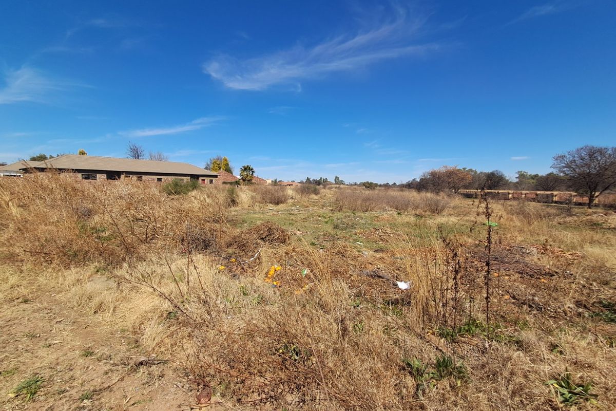 A view of an undeveloped residential plot featuring dry, overgrown vegetation and surrounding neighborhood structures in the distance.