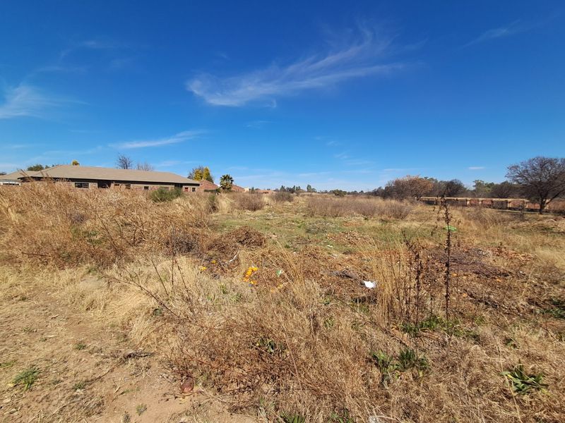 A view of an undeveloped residential plot featuring dry, overgrown vegetation and surrounding neighborhood structures in the distance.
