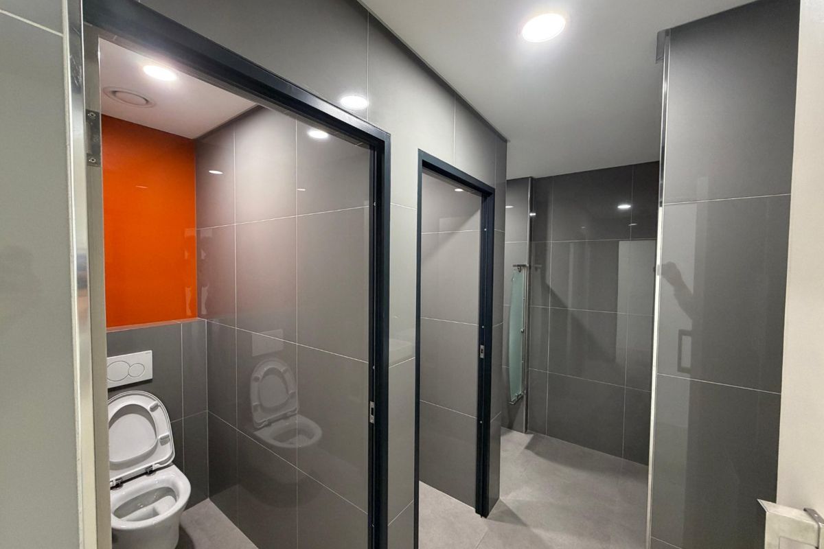 Modern commercial restroom facility featuring grey tiled walls, recessed lighting, and partitioned toilet cubicles with a contemporary orange accent wall.