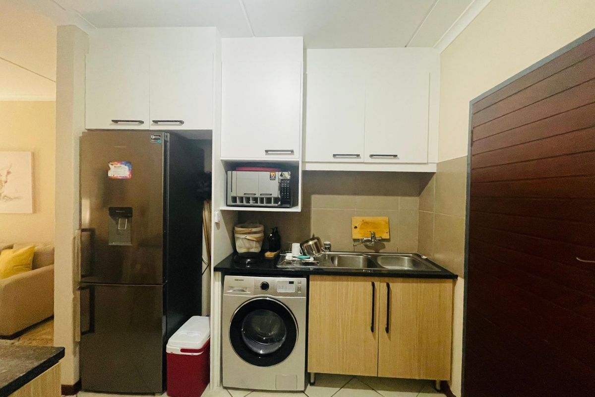 Compact kitchen area featuring white upper cabinetry, wood-finish lower cabinets, a double sink, and tiled backsplash with integrated appliance space.