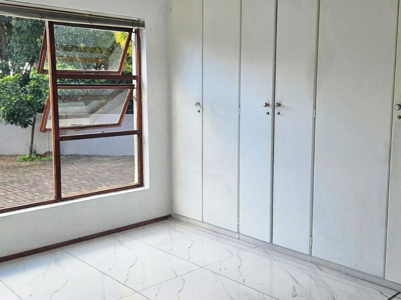 A bright, empty bedroom featuring white built-in cupboards, modern marble-look tiled flooring, and a large window overlooking the paved outdoor area.