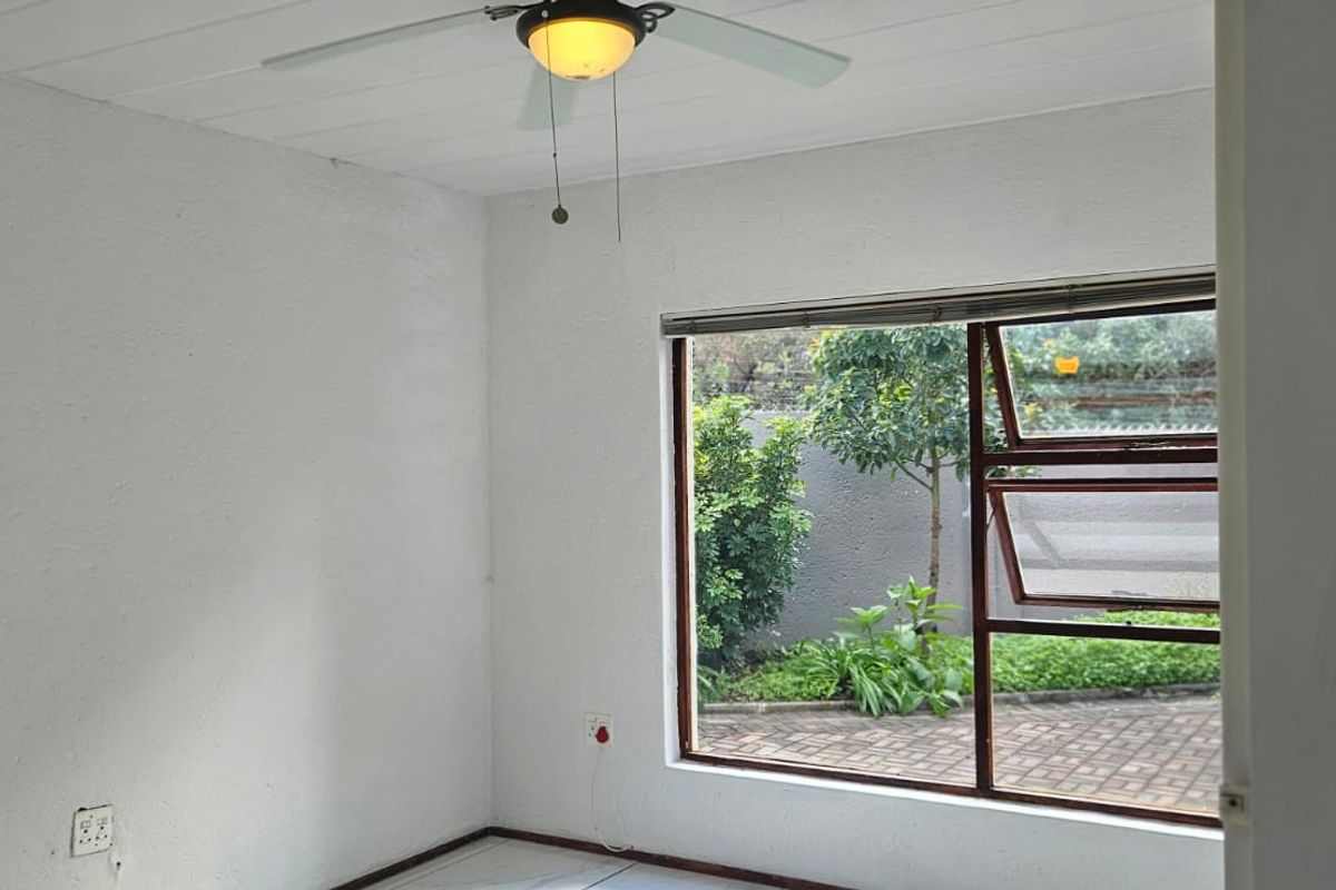 A bright, empty bedroom featuring a ceiling fan and a large window overlooking the garden area.