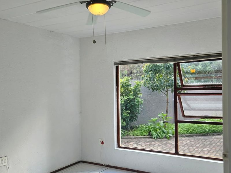 A bright, empty bedroom featuring a ceiling fan and a large window overlooking the garden area.
