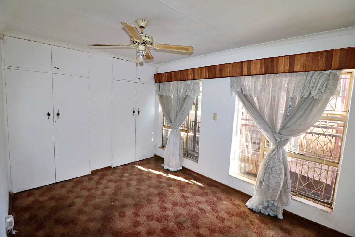 A spacious bedroom featuring built-in cupboards, a ceiling fan, and large windows with security bars, offering a blank canvas for renovation.