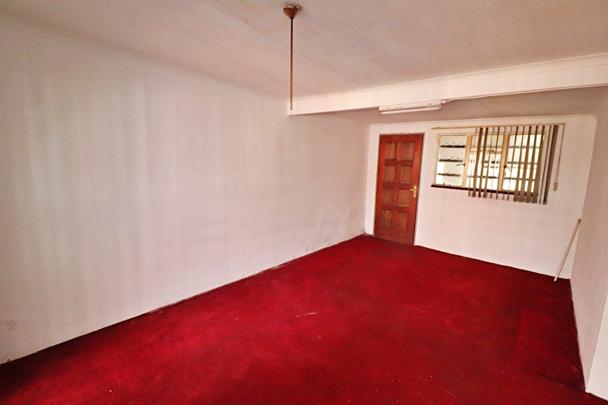 An empty living room featuring red carpet flooring, a timber door, and a window with vertical blinds, requiring cosmetic updates to improve its condition.
