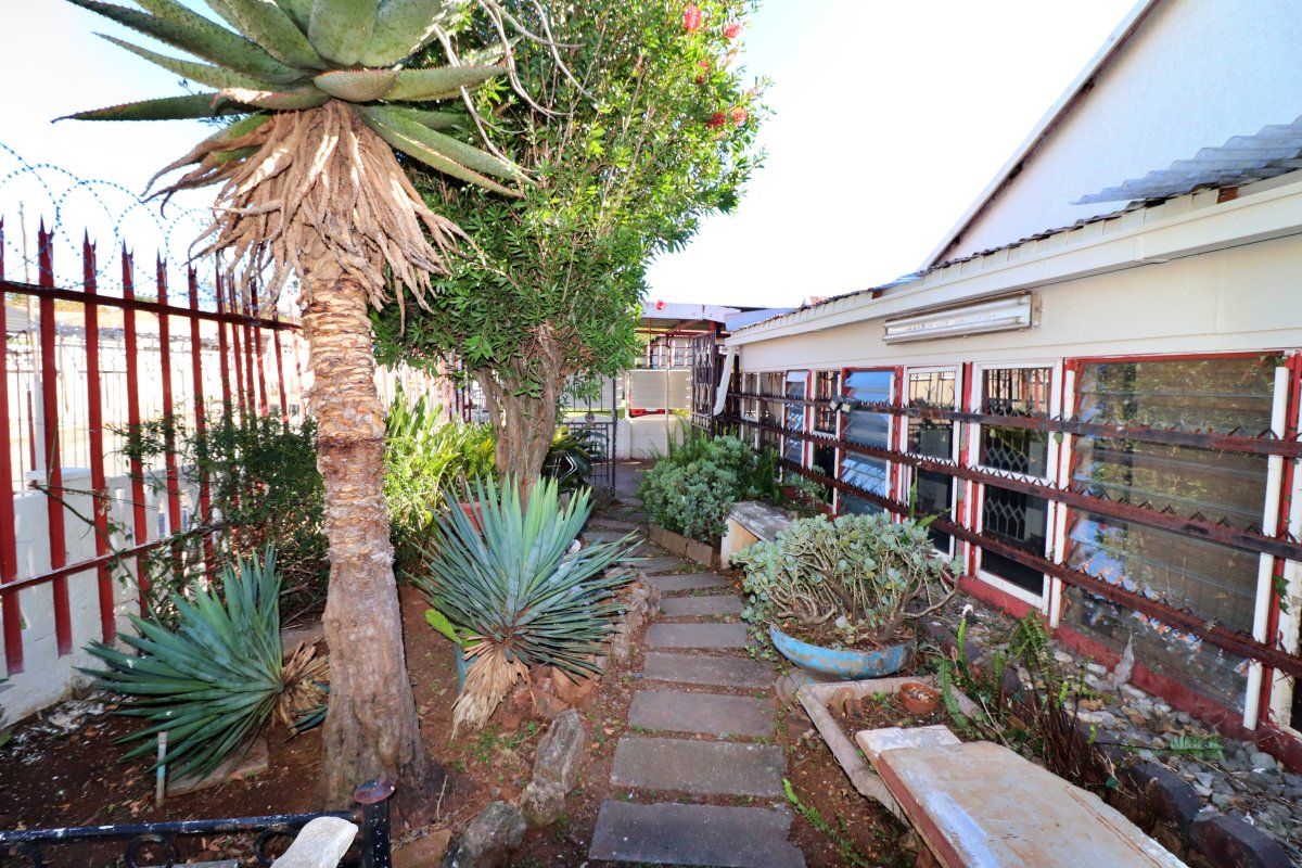 A narrow rear garden pathway featuring established landscaping, security fencing, and access to the side of the house with louvre windows.