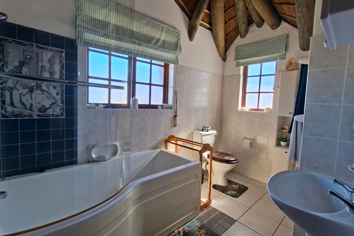 A characterful bathroom featuring exposed timber beams, a thatch ceiling, a modern bathtub with shower screen, and tiled finishes throughout.