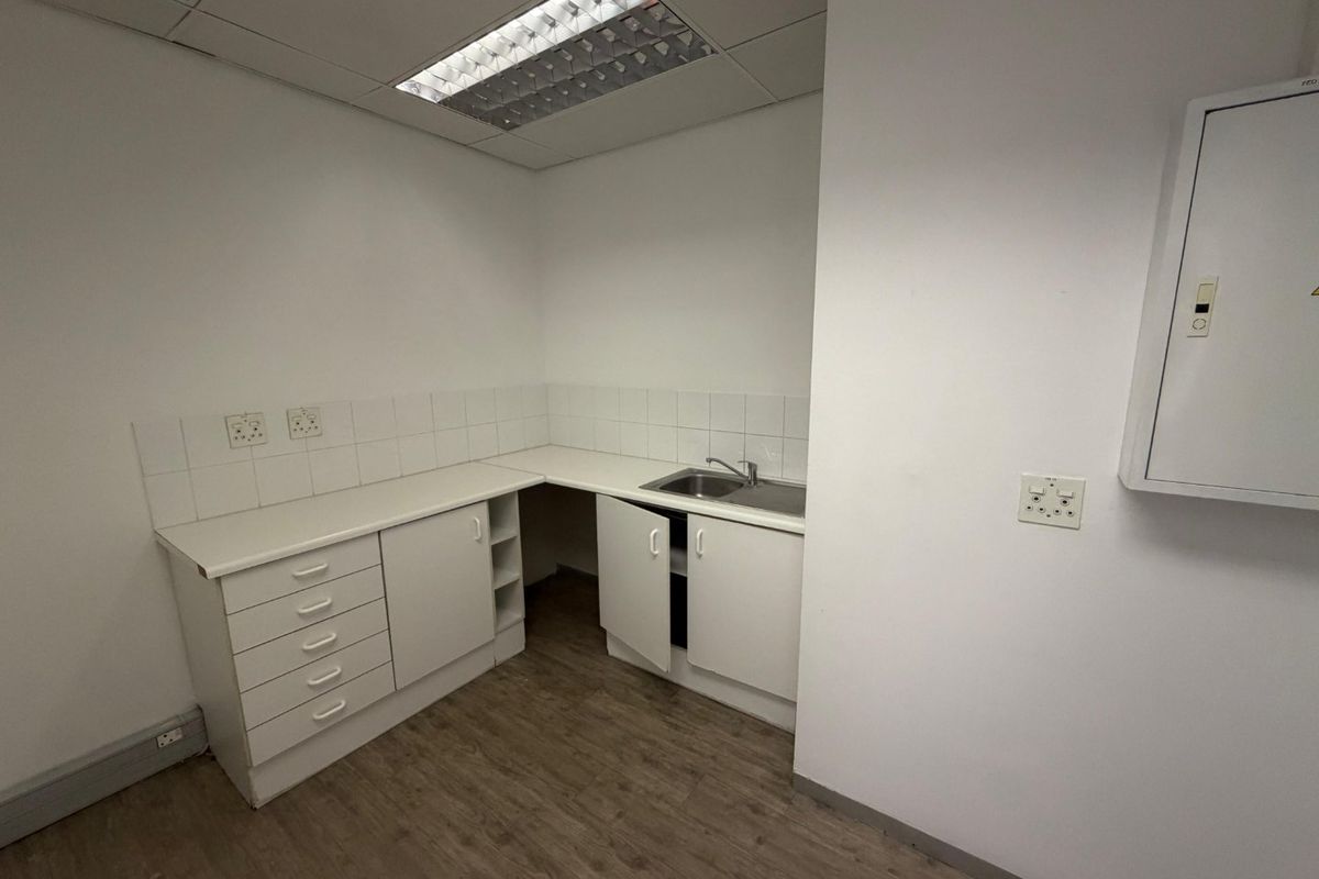 A functional kitchenette area featuring white laminate cabinetry, a stainless steel sink, tiled splashback, and a drop ceiling with integrated lighting.